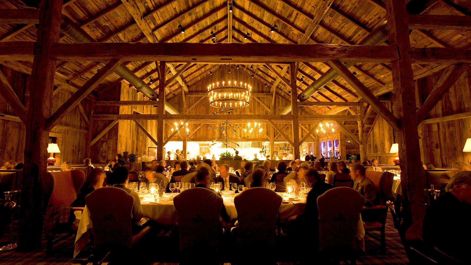 People dining at Blackberry Farm, with a view into the kitchen in the background, exposed wooden beams and rafters along he ceiling, and round chandeliers hanging above